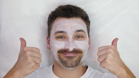 Man with a white mask on his face shows the thumbs of both hands up, nods his head, closes eyes and smiles. Happy facial expressions of male, top view
