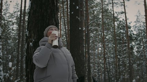 Mature woman in winter clothes resting and drinking tea from thermos cup in forest during her nordic walking
