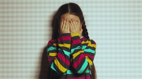 A beautiful dark-skinned little girl in a bright striped sweatshirt joyfully opens and covers her face with her palms. Happy emotional child.