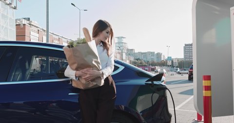 Beautiful Business Woman Stands Near Her Electric Car With Groceries. Electrical Car Charging. Environmentally Friendly Transport Completing Automatic Charging. High-speed Charging Station Near