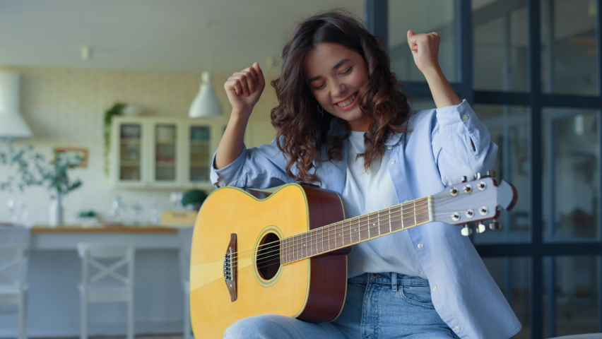 Cheerful girl playing guitar at home. Happy woman practicing music on string instrument in living room. Satisfied guitarist celebrating success. Young lady showing yes gesture with hands | Shutterstock HD Video #1065977764