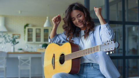 Cheerful girl playing guitar at home. Happy woman practicing music on string instrument in living room. Satisfied guitarist celebrating success. Young lady showing yes gesture with hands