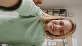 Vertical shot of young smiling caucasian woman showing and talking about polo neck sweater via online video call on smartphone while speaking with customer during workday in clothes shop