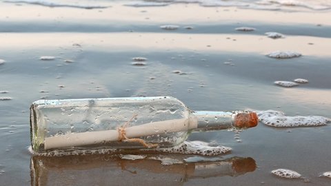 Closeup view of a message in a bottle lying on a wet sandy beach. Waves with foam come towards the bottle from the ocean. Bottle is horizontal with a cork in the opening.