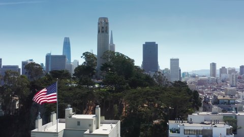 American flag proudly waving with famous San Francisco city on background. Historic Coit tower landmark on hill with cinematic modern financial downtown skyscrapers view. California travel aerial 4K