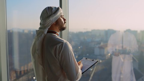 Young Arabic Businessman Reading Papers for Investment Looking out of Window at City Construction. Arab Entrepreneur. Kandura Clothes. Muslim.