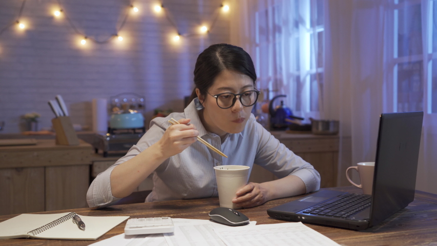 asian chinese businesswoman eating food and sitting at table in home kitchen while looking at computer screen.