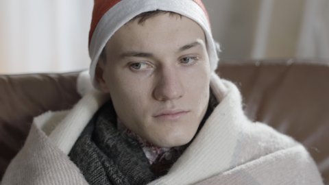 Close-up of depressed young man in Christmas hat sitting on couch at home thinking. Portrait of desperate frustrated lonely Caucasian guy spending Christmas alone indoors. Sadness and loneliness.