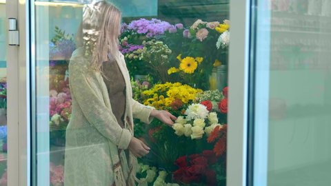 Professional blonde flower shop assistant in long jacket cares and rearranges wonderful bright blooming flowers slow motion