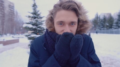 A man warms his hands with warm breath and puts on a hood while standing outside in winter. Close-up. Walking outdoors