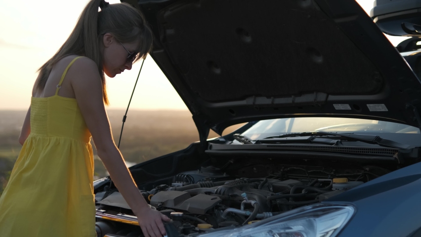 Young puzzled woman driver standing near her car with popped up hood looking at broken engine.