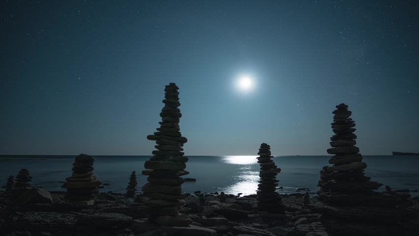 Dolly sideways scenic night many cairn, stones pyramids on shore of calm costal sea, starry sky is moving in circle hyperlapse. Bright full moon light shine through rocks. Abstract seascape, nature 