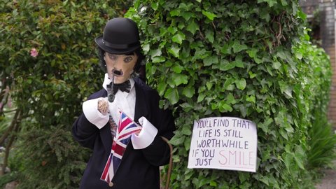 Covid-19 Scarecrow In Charlie Chaplin Costume With A Smoke Pipe And U.K Flag Standing In A Lush Green Garden - Mid Shot