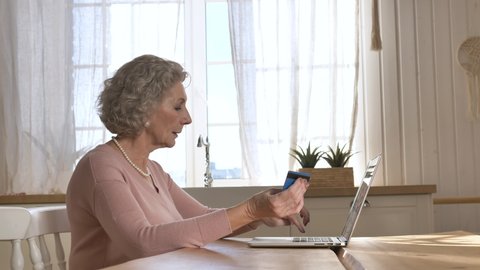 Concentrated elderly woman holds credit card and enters bank information for online shopping sitting at laptop in kitchen closeup