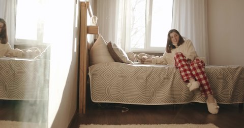 An attractive young girl in a white sweater and bright checkered pants sits on the bed and calls her dog waving a toy