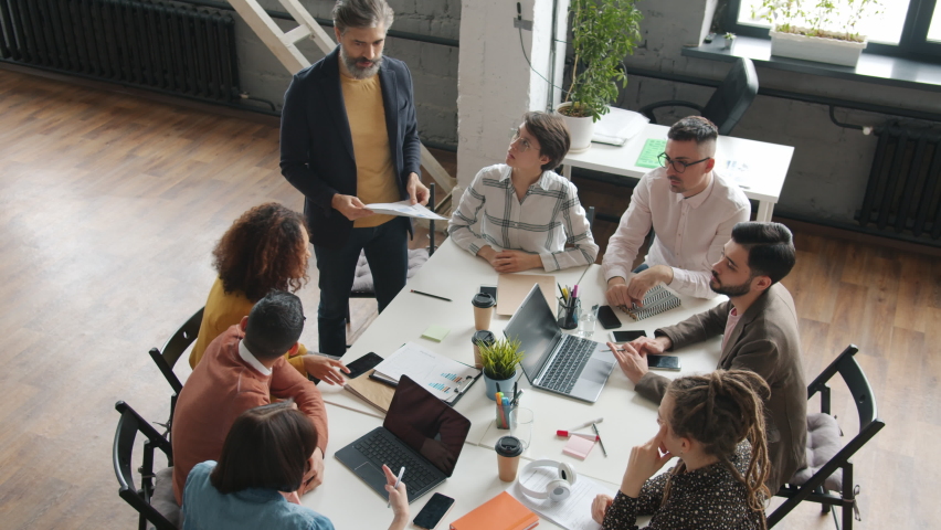 High angle view of creative business team discussing smartphone applications design while mature man distributing papers with device screen in office