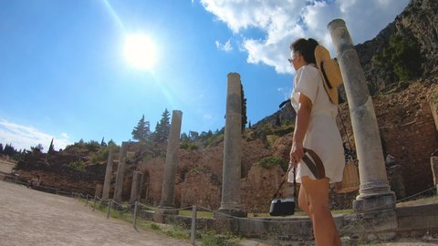 Young woman visiting ruins of temple of Apollo in Delphi, archaeological site by mount Parnassus, Greece, Europe. Large hat, fashion white dress.