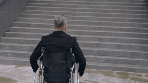 Crop view of disabled male person in suit pushing himself on wheel chair at street. Disabled man going to work and stopping near office building stairs. Concept of obstacles,barriers