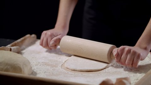 Closeup Shot Of Female Hands Of Baker Rolls Dough In Flour On Wooden Board On Table With A Rolling Pin, In Bakery Shop, Woman Making Pizza Using Traditional Recipe At Home On A Black Background