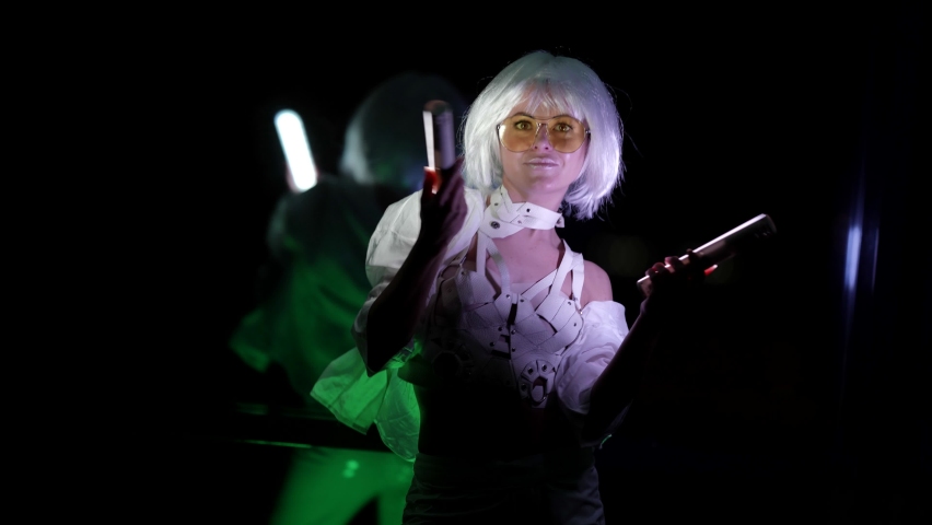 A beautiful blonde woman in stylish glasses in a white sexy suit is dancing in the studio against a dark background in front of a mirror, holding two LED lights in her hands.