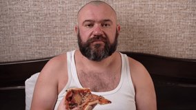 a bearded man sits on the bed at home, makes a video call via a webcam and eats pizza. Communicates with a mouth full