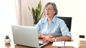 Concentrated senior businesswoman has virtual meeting on a laptop, sitting at the desk talking, checking notes, middle-aged lady boss distributes remotely errands, tasks in team via video call