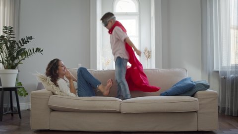 Mom And Son In The Room. Boy In Red Cloak And Glasses Plays. He Jumps Up To His Mother On The Sofa. They Have Fun. Beautiful Interior In Light Colors.