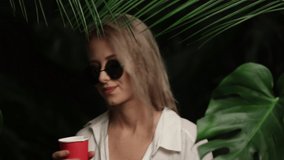 Blonde woman dancing and drinking near palm leaves