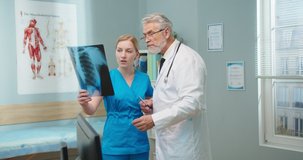 Adult Caucasian male doctor talking to pretty female murse holding x-ray of lungs. Young assistant listening to senior man pointing to scan and explaining disease in hospital. Healthcare concept.