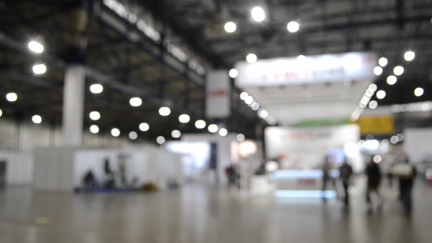 Abstract blurred background. Inside big building. People walk inside space. Lots of glowing spotlights, lamps shine indoors. Beige floor, black-gray ceiling. Many stands and equipment for exhibitions