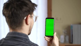 View over shoulder on vertical smartphone with green screen in hands of caucasian man. Guy sitting at table outdoor. Chroma key. Male video chat on webcam of mobile phone.
