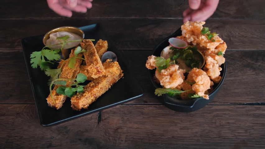 Two hands serving two prepared dishes on a wooden surface. Crispy seafood fried crab dishes with parsley. Slowmotion slow tilt-up