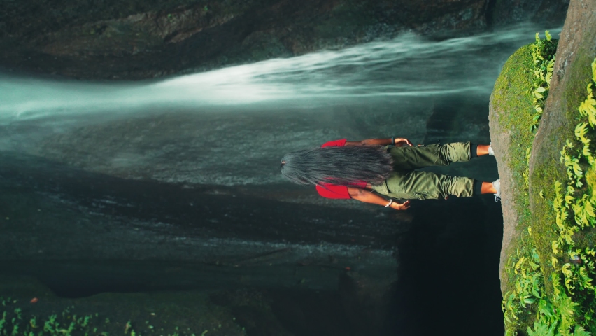 Young attractive girl explores the picturesque waterfall hidden in tropical rain forest jungle. 4K