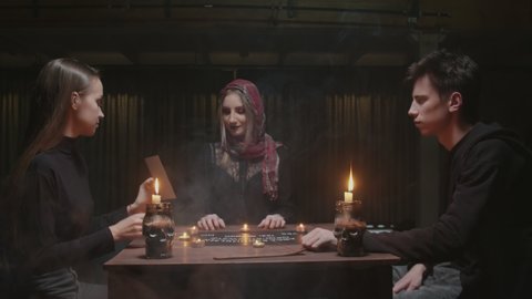 Young couple visits young witch female fortune teller. Young woman shows photo of her lost friend to the witch, then they takes each others hands and closes eyes