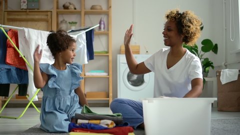 Young american woman and little girl having fun and sitting on floor in laundry room at home spbd. Cheerful mother and cute girl clap their hands and laugh, sit in bright interior with washing machine