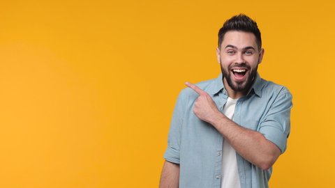 Excited funny bearded young man 20s years old in casual basic blue shirt isolated on yellow color background studio. People lifestyle concept. Pointing index finger aside showing thumb up like gesture