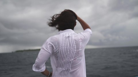 The woman stands with her back to the camera and looks into the distance at the raging storm on the sea and dark gray clouds and the ship sways in different directions.