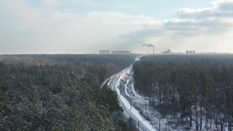 Flying over railway in the winter forest on industrial city background.