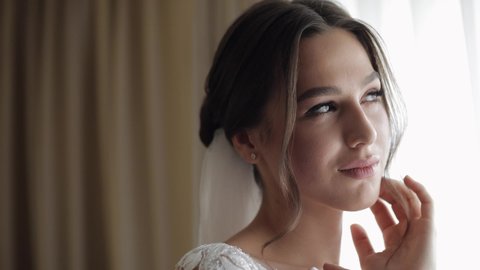Bride in white dress staying near window and touching her face. Wedding morning preparations. Bride with fashion hairstyle and makeup in hotel or home apartment. Beautiful, sexy woman in gown and veil