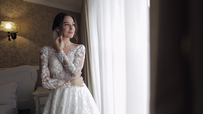 Bride in white wedding dress stay near window. Wedding morning preparations. Luxury bride with fashion hairstyle and makeup in a hotel or apartment. Beautiful, lovely woman in dress and veil