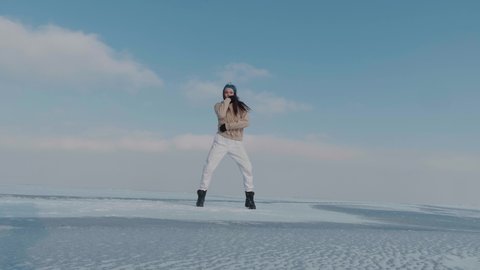 Stylish young woman, professional dancer, dancing energetic jazz funk or hip hop on frozen lake in cold winter. Female hipster dances challenge for social networks on snow-covered frozen arctic river