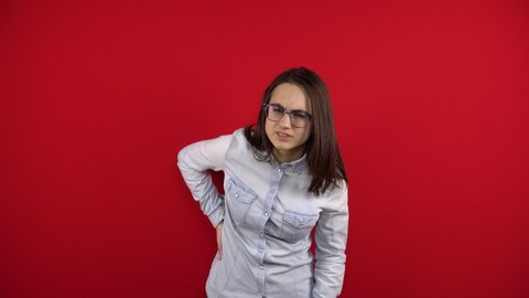 A young woman with glasses has a backache and is holding it in her hands. Shooting on a red background.