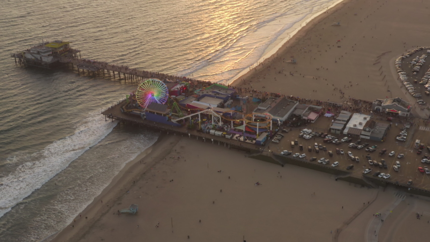 Flying towards Santa Monica Pier, Los Angeles at beautiful Sunset with Tourists, Pedestrians having fun at Ferris Wheel with ocean view waves crashing, Aerial Drone Perspective Royalty-Free Stock Footage #1066078858