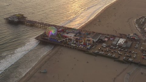 Flying towards Santa Monica Pier, Los Angeles at beautiful Sunset with Tourists, Pedestrians having fun at Ferris Wheel with ocean view waves crashing, Aerial Drone Perspective