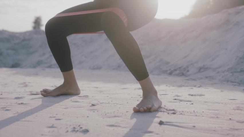 Close up african american woman in sportswear doing squats with resistance band during workout on sunny day on sand desert dunes. Muscular practice stretching squatting sportswoman outdoor. Slow
