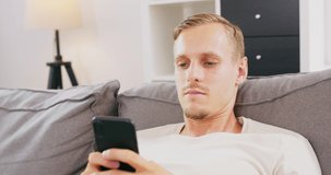 Man lying relaxing on comfortable sofa at home chatting texting on smartphone, young caucasian european male rest on comfy couch use cellphone, messaging or browsing internet on cell.