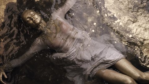 a woman in a dress with dyed gold skin and hair lies in the water, slowly moving. She strokes her body.