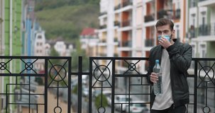 Young handsome man takes off his medical protective face mask and drinks mineral water from big plastic bottle. Cinema 4K panoramic slow motion video