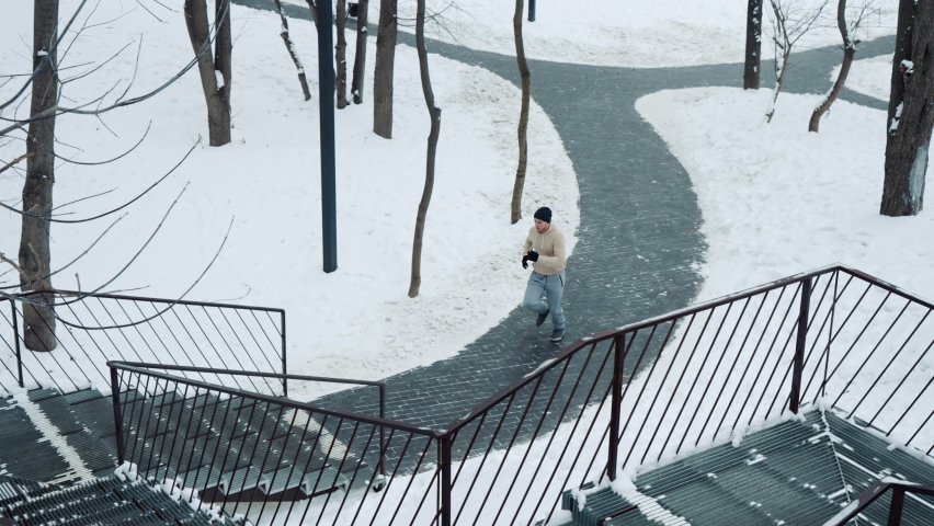 Fit man in sportswear, hat and gloves running up on metal staircase and approaching camera, snow lying on ground. Slow motion athlete jogging in city park in winter. Concept of sport