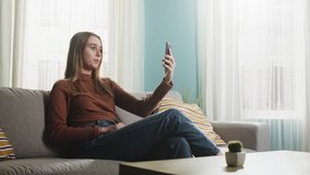 A young lady in brown sweater and blue jeans sits on the sofa in front of window, talking on video and saying goodbye to her interlocutor. The average plan